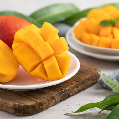 Ahşap kesim tahtasının üzerinde beyaz bir tabakta kesilmiş taze mango meyvesi ve gri masa arkasında yapraklar..