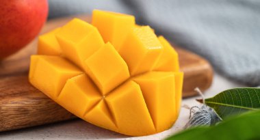 Ahşap kesim tahtasının üzerinde beyaz bir tabakta kesilmiş taze mango meyvesi ve gri masa arkasında yapraklar..