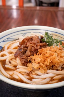 Biftek, soğan ve çıtır tempura kırıntılarıyla kaplı bir kase Japon körili udon eriştesi..
