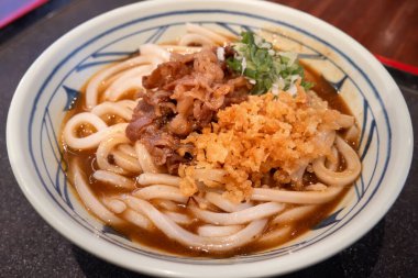 Biftek, soğan ve çıtır tempura kırıntılarıyla kaplı bir kase Japon körili udon eriştesi..