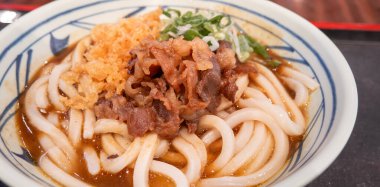 Biftek, soğan ve çıtır tempura kırıntılarıyla kaplı bir kase Japon körili udon eriştesi..