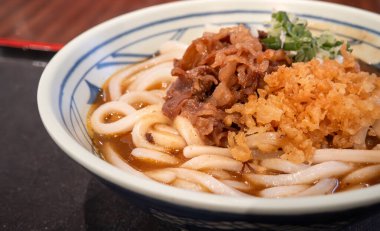 Biftek, soğan ve çıtır tempura kırıntılarıyla kaplı bir kase Japon körili udon eriştesi..