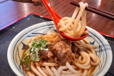 Biftek, soğan ve çıtır tempura kırıntılarıyla kaplı bir kase Japon körili udon eriştesi..