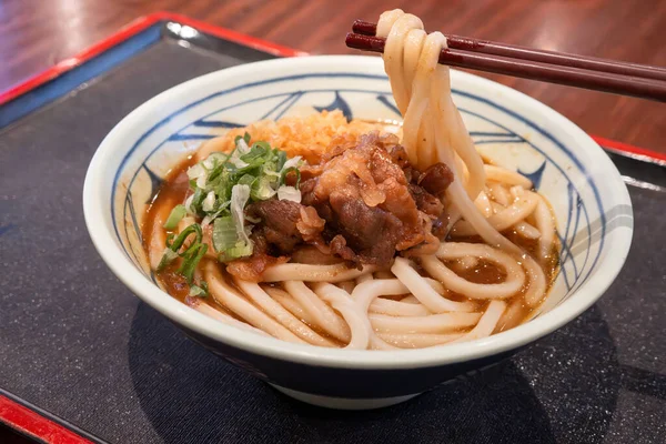 Biftek, soğan ve çıtır tempura kırıntılarıyla kaplı bir kase Japon körili udon eriştesi..