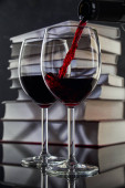 Červené víno se nalije do sklenice z láhve na pozadí stohu knih, proud červeného vína z láhve víří ve sklenici, zblízka.