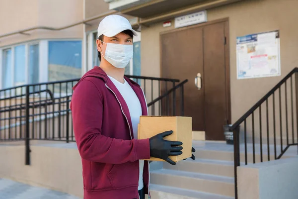 Tıbbi lastik eldivenli ve koruyucu maskeli bir kurye karton kutu tutuyor. Teslimat servisi. Karantina.