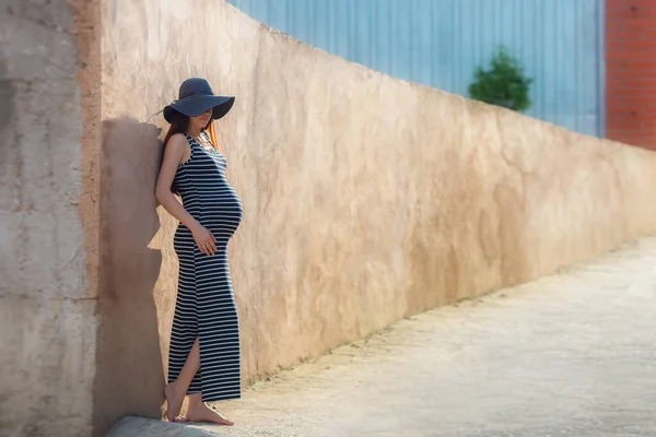 Tarafsız arka planda şapkası olan siyah elbiseli güzel, şık hamile bir kadın..