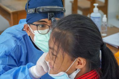 BALI / INDONESIA-MAY 28 2021: Bir ENT doktoru bir pediyatrik hastanın kulaklarını, burnunu ve boğazını muayene ediyor. COVID-19 salgını sırasında, ENT incelemeleri çok risklidir çünkü virüsün bulunduğu organlardaki hastalarla doğrudan temasta bulunurlar. 