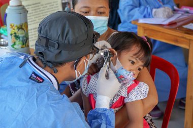 BALI / INDONESIA-MAY 28 2021: Bir ENT doktoru bir pediyatrik hastanın kulaklarını, burnunu ve boğazını muayene ediyor. COVID-19 salgını sırasında, ENT incelemeleri çok risklidir çünkü virüsün bulunduğu organlardaki hastalarla doğrudan temasta bulunurlar. 