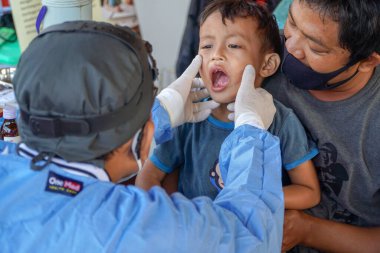 BALI / INDONESIA-MAY 28 2021: Bir ENT doktoru bir pediyatrik hastanın kulaklarını, burnunu ve boğazını muayene ediyor. COVID-19 salgını sırasında, ENT incelemeleri çok risklidir çünkü virüsün bulunduğu organlardaki hastalarla doğrudan temasta bulunurlar. 