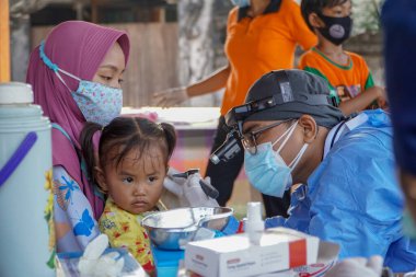 BALI / INDONESIA-MAY 28 2021: Bir ENT doktoru bir pediyatrik hastanın kulaklarını, burnunu ve boğazını muayene ediyor. COVID-19 salgını sırasında, ENT incelemeleri çok risklidir çünkü virüsün bulunduğu organlardaki hastalarla doğrudan temasta bulunurlar. 
