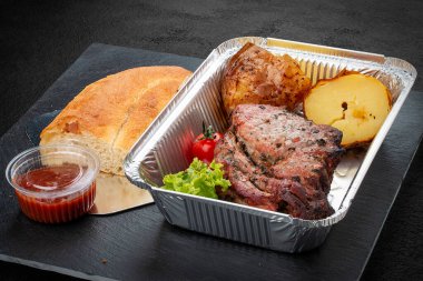 Fırında patates, kibrit ve kırmızı soslu ızgara domuz eti. Yemek servisi. Alüminyum beslenme çantasında servis edilmiş.. 