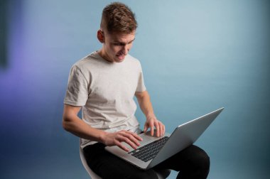 Genç adam mavi arka planda dizüstü bilgisayar kullanıyor. Genç, beyaz bir adam dizüstü bilgisayarla çalışıyor. Sandalyeye oturmuş dizüstü bilgisayarla uğraşıyor.