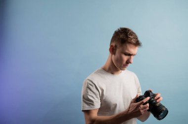 Mavi arka planda kamera kullanan yakışıklı genç bir fotoğrafçı. Profesyonel kamera kullanan bir adam. Fotoğraf makinesine bakan bir adamın portresi.