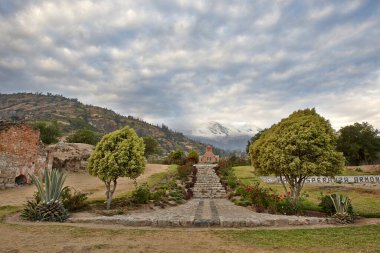 Arka planda karlı dağlar olan küçük bir kiliseye çıkan merdivenli bir bahçe. Ağaçlar ve kırmızı güller var.