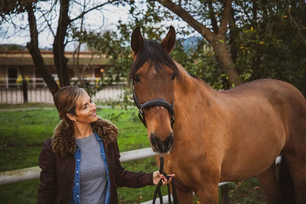 Wesoła Uśmiechnięta Kobieta Ubrana Brązową Kurtkę Patrząca Swojego Konia Ranczu — Zdjęcie stockowe