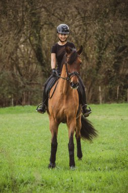 Kahverengi atıyla siyah giyinmiş kadın binici ağaçlarla dolu yeşil bir çayırda at sürüyor.