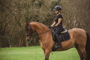 Kahverengi atıyla siyah giyinmiş kadın binici ağaçlarla dolu yeşil bir çayırda at sürüyor.