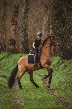 Kadın binici, siyah giyinmiş, kahverengi atıyla kır yolunda gidiyor.