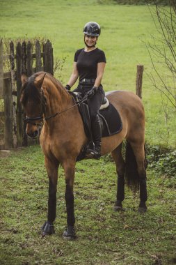 Kırsal kesimdeki bir çitin önünde siyah toynaklı siyah giysili kadın binici gülümsüyor.