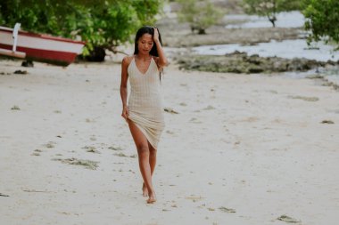 Çıplak ayaklı bir kadın, beyaz bir kumsalda rahat bir yaz elbisesiyle yürüyor.