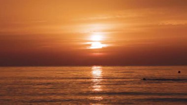 Şafak vakti açık denizde tek başına yüzen bir insanın silueti. Akdeniz kıyısında sıcak gündoğumu tonları ve nazik dalgalarla çevrili.