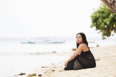 Sakin bir kıyı şeridinde poz veren rahat bir kadın. Uzakta küçük tekneler, yalnızlık, yansıma ve huzurlu bir tropikal yaşam tarzı..