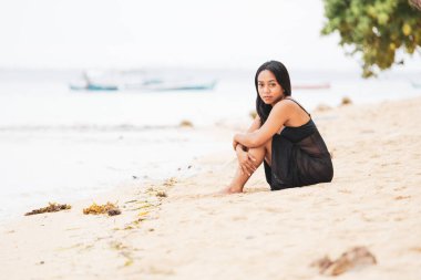 Sakin bir kıyı şeridinde poz veren rahat bir kadın. Uzakta küçük tekneler, yalnızlık, yansıma ve huzurlu bir tropikal yaşam tarzı..