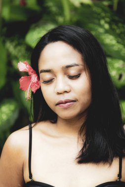 Doğal güzellik portresi. Canlı yeşil yapraklar arasında poz veren bir kadın. Kulağının arkasına saklanmış kırmızı bir çiçek, dinginlik, tropikal zarafet ve doğa ile olan bağlantısını ifade ediyor..