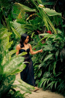 Etrafı yoğun yeşil yapraklarla çevrili zarif bir kadın, canlı bir orman yolunu incelerken nazikçe yapraklara dokunuyor, kendine güvenini, özgürlüğünü ve huzurlu bir yolculuk yaşam tarzını ifade ediyor..