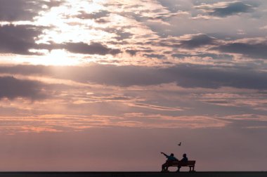 İki kişi bir bankta oturmuş dramatik günbatımını izliyorlar. 