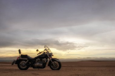 Canlı bir günbatımı gökyüzünün altında park etmiş bir motosiklet, özgürlük, macera ve bağımsızlığı çağrıştırıyor, ve açık yolların ruhunu yansıtıyor, kişisel keşifler, ve ufkun ötesine geçmek için sonsuz arzu..