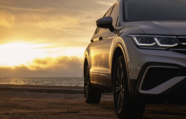 Renkli bir günbatımının gölgesinde gri bir araba okyanusun kenarında duruyor. Sıcak ışık ve açık alan bir yalnızlık ve keşif anını yakalar.