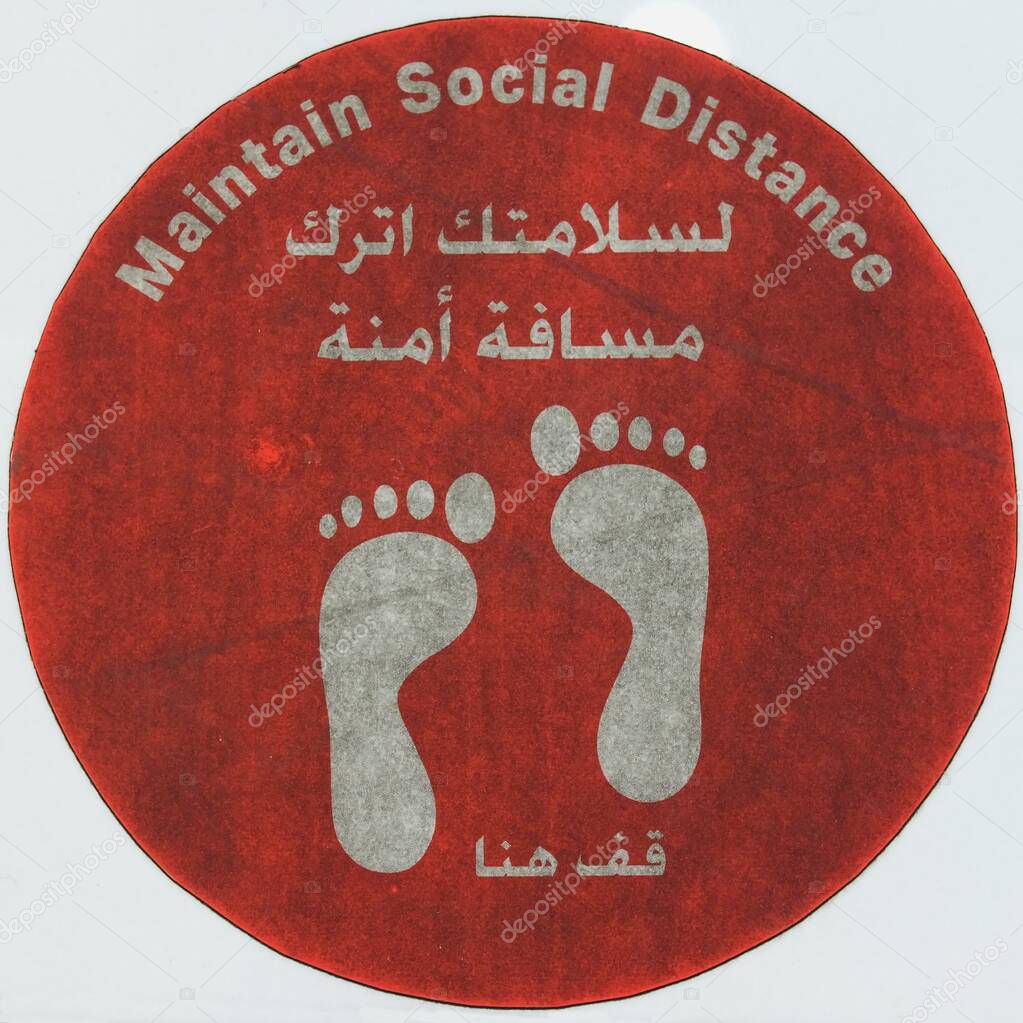 Dirty footworn round red colored sign MAINTAIN SOCIAL DISTANCE, coronavirus disease COVID-19 warning sign written in both in Arabic and English on the floor of shopping mall.
