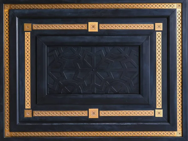 Геометрический дизайн фрагмента современной роскошной черной деревянной входной двери с резными панелями и золотыми рамками из патины с классическим орнаментом. Стоковая Картинка