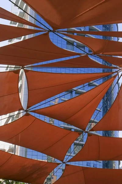 Creativa construcción de dosel al aire libre de toldos triangulares de tela de color naranja oscuro del sol contra rascacielos residenciales modernos. Toldos de protección solar para infraestructuras urbanas. — Foto de Stock