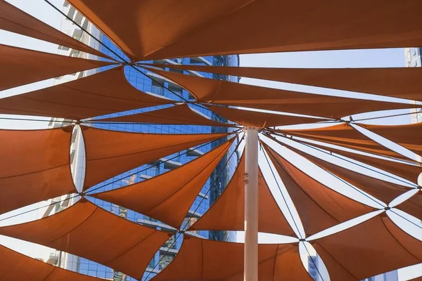 Creativa construcción de dosel al aire libre de toldos triangulares de tela de color naranja oscuro del sol contra rascacielos residenciales modernos. Toldos de protección solar para infraestructuras urbanas. — Foto de Stock