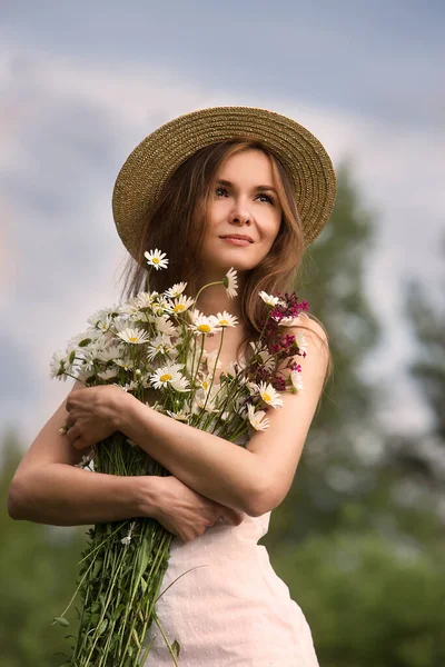 空に対してデイジーと野の花の花束とわらの帽子の若い美しい女性の肖像画 — ストック写真