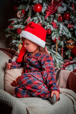 Pijamalı ve Noel Baba şapkalı mutlu bir kız çocuğu Noel ağacının yanında otururken gülümsüyor. Sihir. Yeni yıl arifesi. Aziz Niklas Günü. Hediye. Tatil ve mucizeler zamanı.