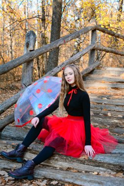 Parktaki ahşap bir köprüde oturan kırmızı etekli, dalgalı sarı saçlı genç bir kızın dikey görüntüsü