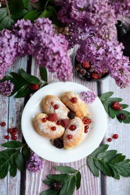 Beyaz bir tabağın üst görüntüsü, arka planda leylak dalları olan küçük kruvasanlarla dolu. Dikey.