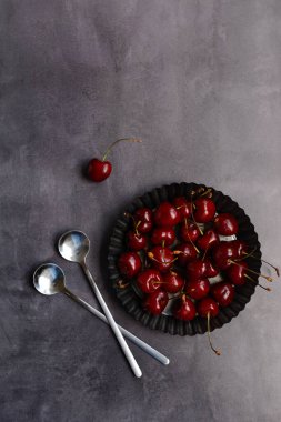 Dikey yerleşim. Bir tabakta tatlı kirazlar ve koyu gri arka planda kaşıklar. Üst görünüm, boşluğu kopyala. Minimalizm
