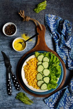 Dikey kompozisyon. Salatalık salatası, mısır, baharat, marul, kaşık, çatal ve peçeteyle mavi arka planda. Lezzetli ve sağlıklı yemek kavramı.