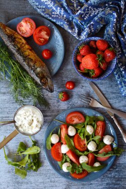Vertical composition. Grilled fish, strawberries, salad with tomatoes, cheese and arugula on a blue background, top view. The concept of tasty and healthy food.