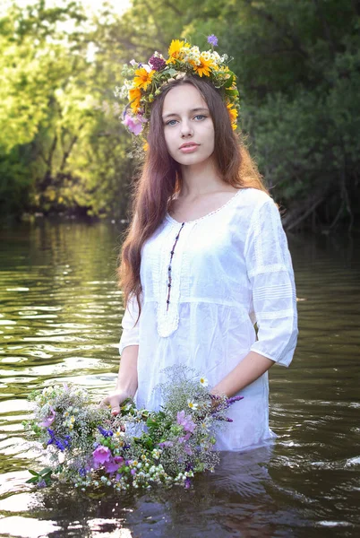 Portrait of a young beautiful girl in white with a wreath on her head against the background of the river. Holiday of Ivan Kupala. Slavic traditions.