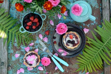 Çay içmenin güzel bir bileşiminin üst görüntüsü. Böğürtlenli kekler, ahududu ve böğürtlen bir tabakta, çaydanlık, çiçekler, turkuaz arka planda gül yaprakları
