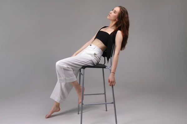 Young woman posing on a chair in the studio in a black top and white pants. Fashion portrait of a young woman on a gray background.