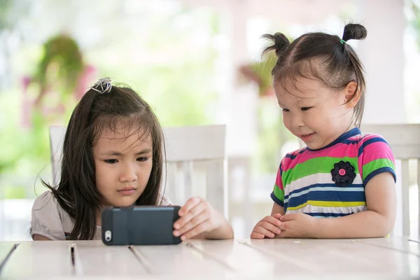 Två lite asiatisk tjej sitter på stol med hjälp av mobiltelefon — Stockfoto