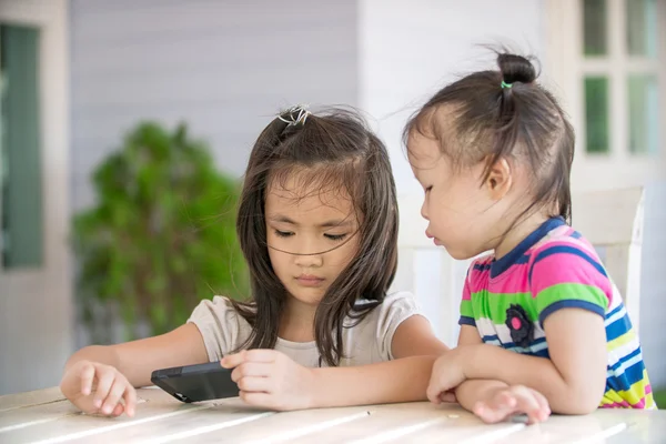 Dvě malé asijské dívka sedí na židli pomocí mobilního telefonu — Stock fotografie