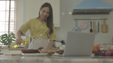 Güzel bir Asyalı kız, mutlu bir yüzle dizüstü bilgisayarıyla nasıl yapılacağına bakarak kahvaltı hazırlar. Kendi evinde.